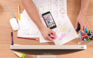 a person seats in front of a desk top with cell phone and documents