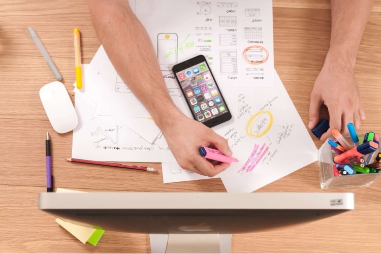 a person seats in front of a desk top with cell phone and documents